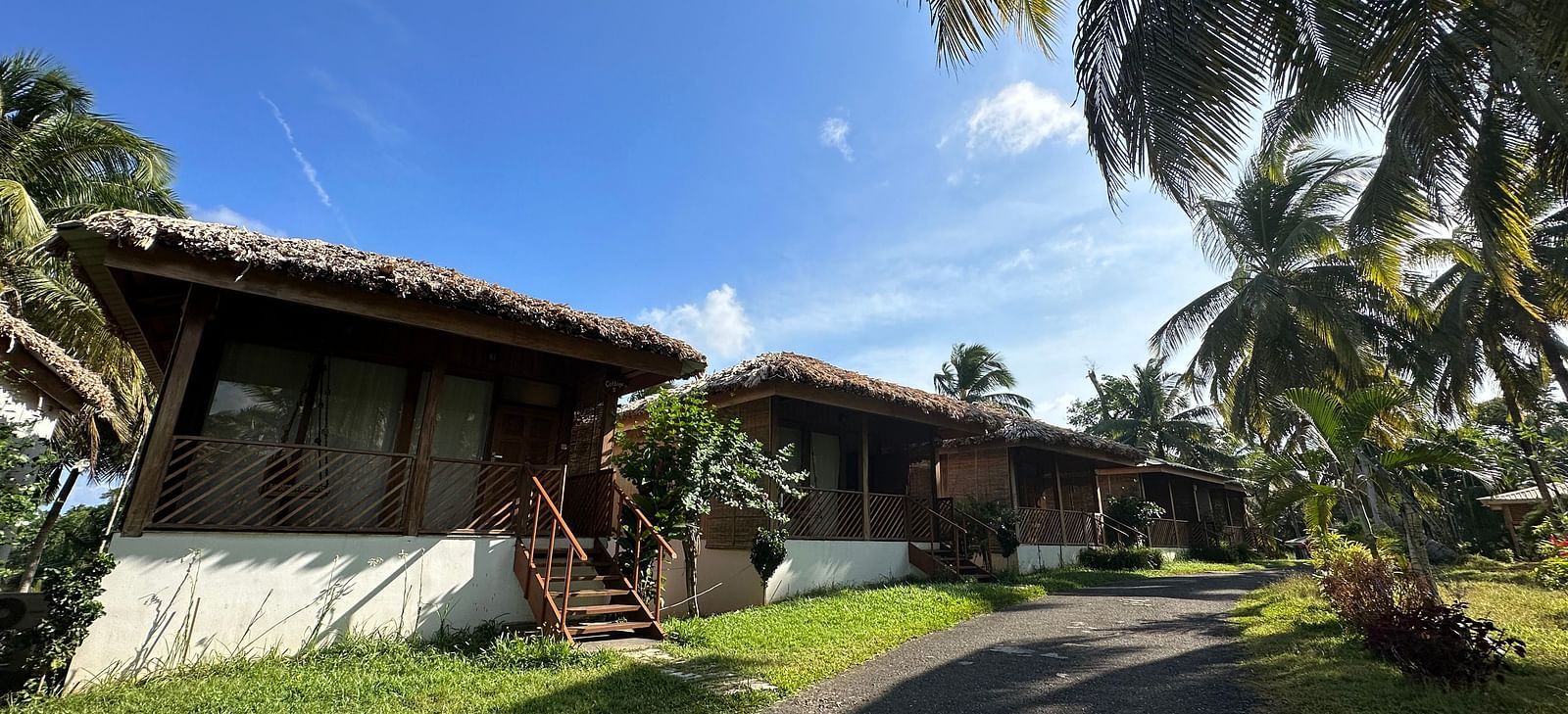 A charming resort building with a thatched roof, surrounded by lush green grass and palm trees under a bright sky.