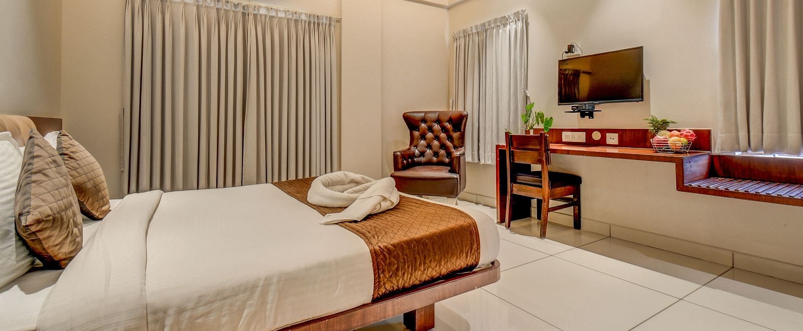 A view of a spacious bed, a wall-mounted TV with a desk beneath it, and a chair next to the side table at Harigivinda's JNStay