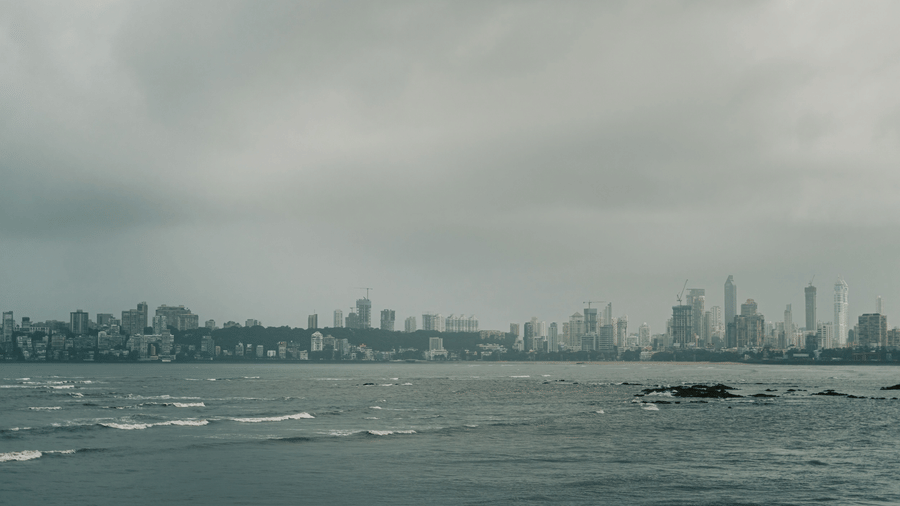 Grey monsoon skies stretch across the Arabian Sea, with Mumbai's skyline visible in the distance.