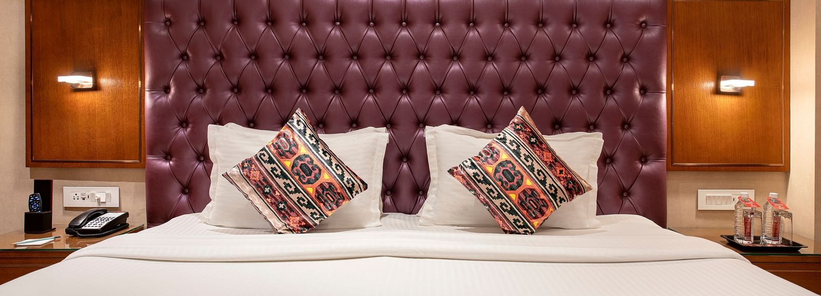 A close-up of a comfortable hotel bed featuring a plush purple headboard and decorative cushions with an ornate bed runner - SK Lords Inn Ahmedabad