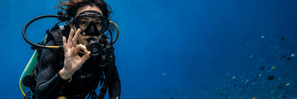 A scuba diver swimming underwater above a coral reef in clear blue ocean water.