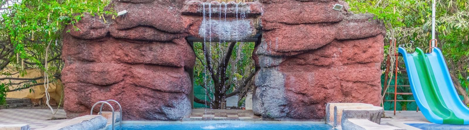 A swimming pool with a slide featuring a man-made waterfall at the background at Daksh Resort & Amusement Park.
