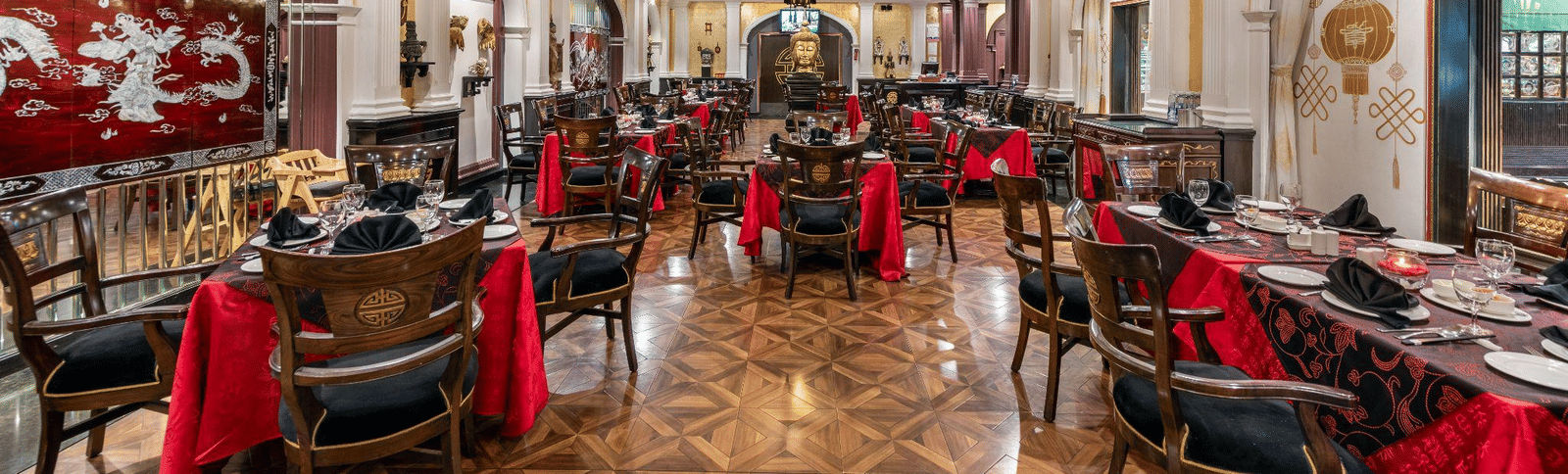 Grand banquet hall or wedding reception venue with a long, vaulted ceiling, elaborate chandeliers, and tables dressed in red and black accents.