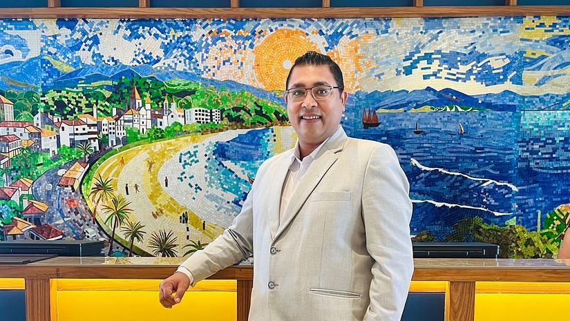 A person wearing a light-colored suit stands in front of a brightly colored, abstract painting with geometric patterns.