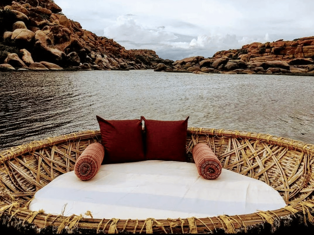 A round, woven coracle boat, styled with a comfortable white mattress and two deep red bolster pillows, floats on a serene river.