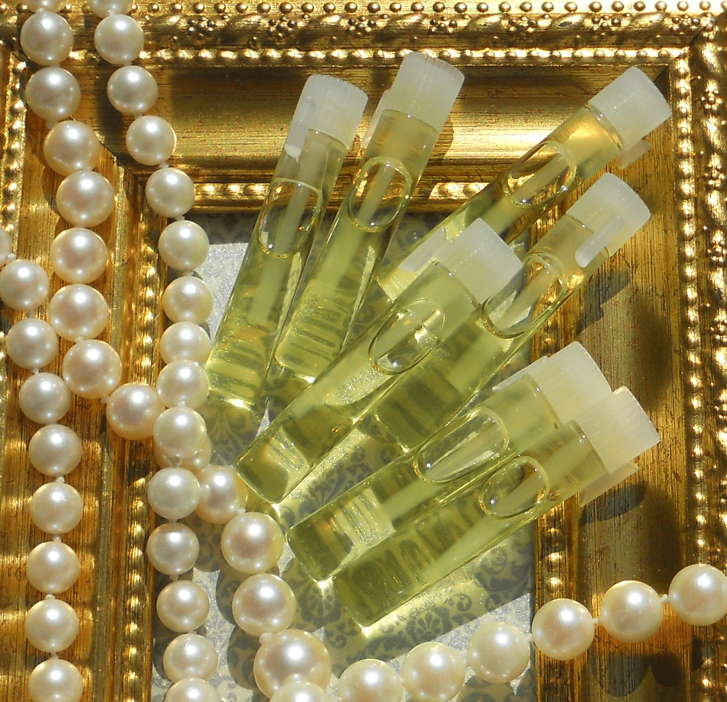 The image shows several small bottles of  oil arranged in a decorative frame. There are pearls surrounding the frame. The bottles appear to be filled with a clear, yellow liquid.
