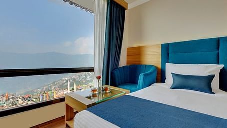 An image of the premium executive room with a bed, a table, a chair and an expansive window overlooking the view - Yashshree Sikkim Blossom, Gangtok