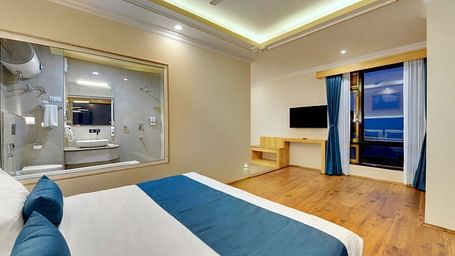 An image of the premium executive room with a bed, flat screen TV, a window overlooking the view and the washroom area is visible through an expansive window - Yashshree Sikkim Blossom, Gangtok