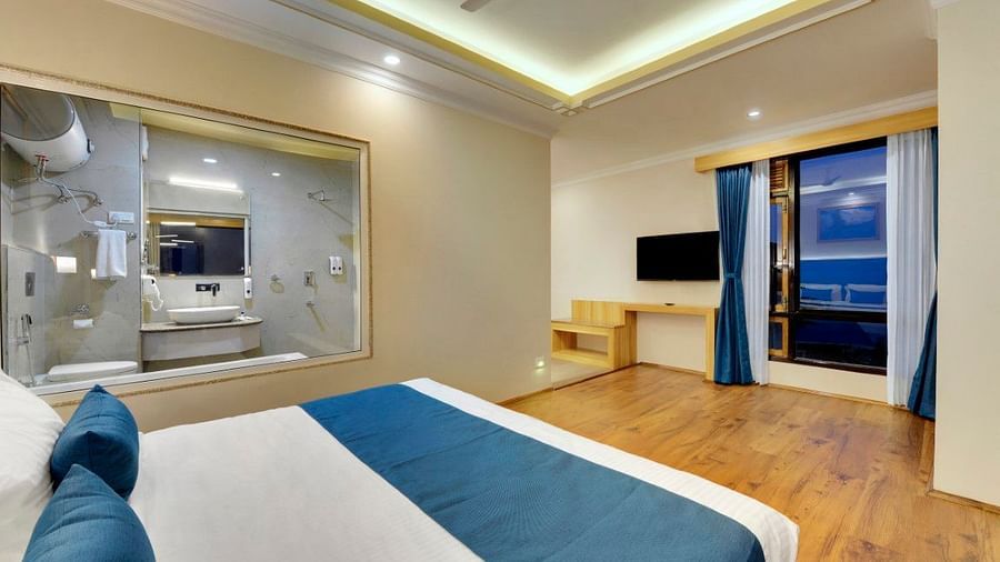 An image of the premium executive room with a bed, flat screen TV, a window overlooking the view and the washroom area is visible through an expansive window - Yashshree Sikkim Blossom, Gangtok