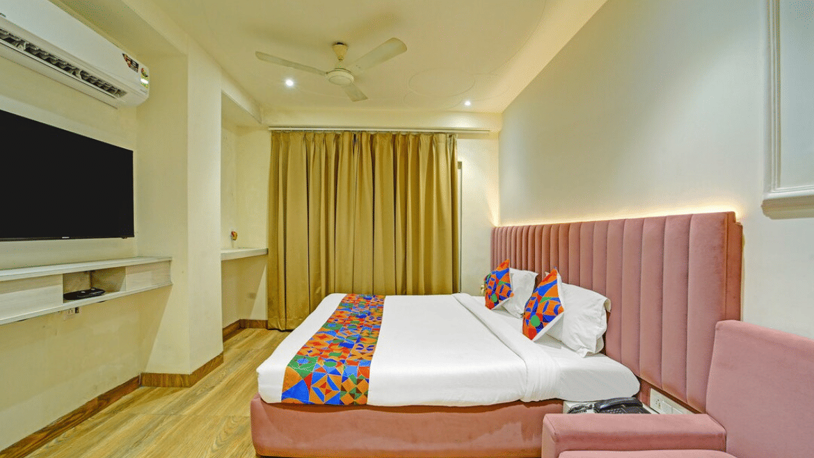 Bedroom with double bed, colorful runner and cushions, pink sofa, wall-mounted TV and curtains at Hotel Maurya, Bhopal.