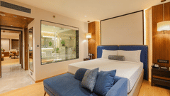 Contemporary bedroom at Pride Premier Pune with a king size bed, blue seating and a glass partition to the bathroom.