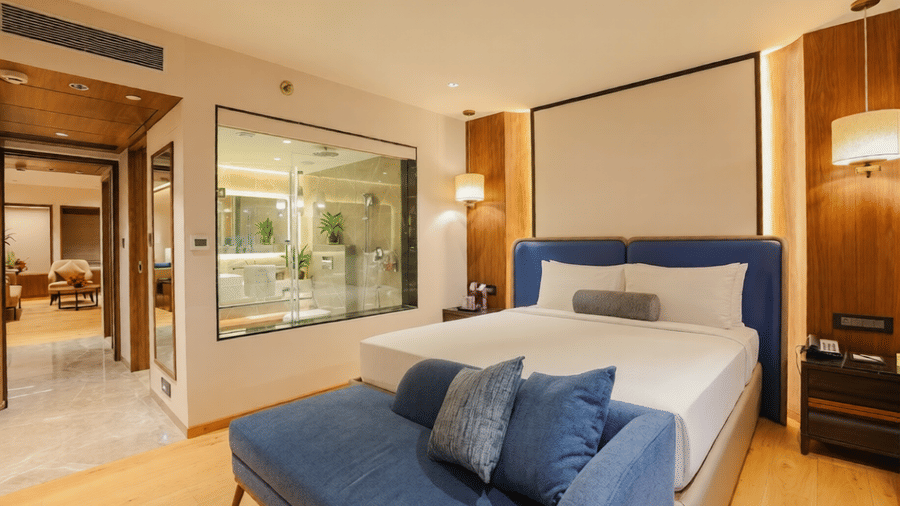 Contemporary bedroom at Pride Premier Pune with a king size bed, blue seating and a glass partition to the bathroom.