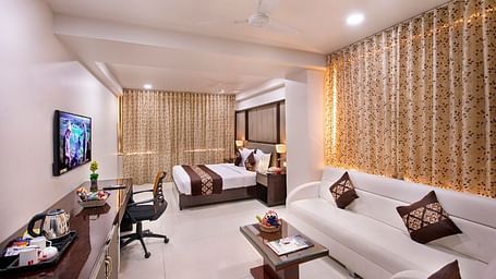 A well-appointed hotel room features two neatly made single beds, a work desk with a chair, and a wall-mounted television.