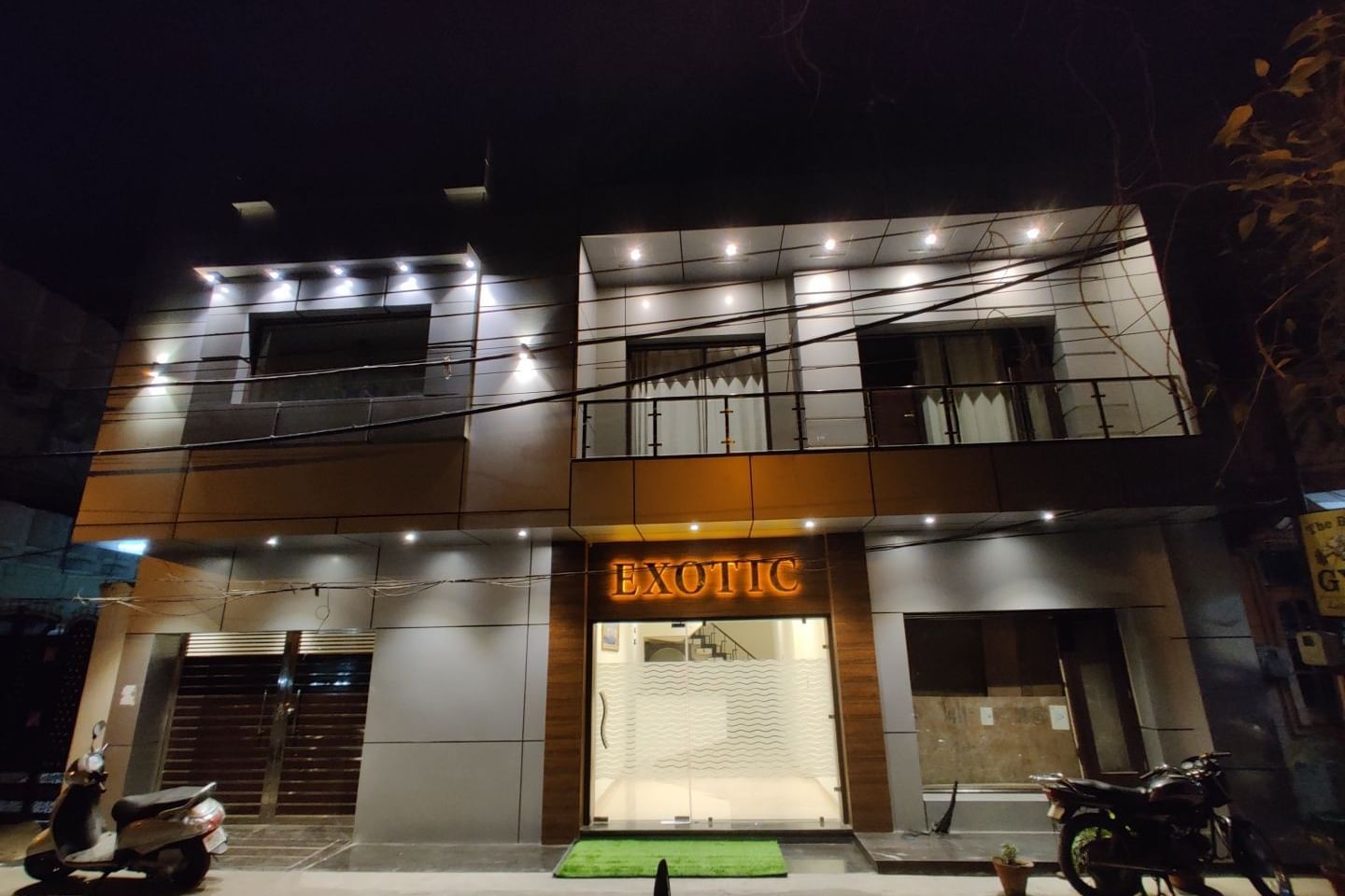 An exterior shot of Hotel Exotic Amritsar at night, showing the illuminated signage and the modern facade of the building.