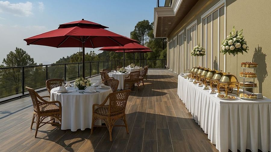  Al fresco buffet and dining setup on a sunny terrace at The Manor Sports & Wellness Hotel, with red umbrellas, white linen tables and views of pine-covered hills.