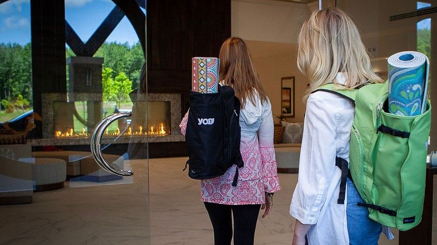 Two women with backpacks and yoga mats walking into YO1 Longevity and Health Resorts with large windows, elegant decor, 