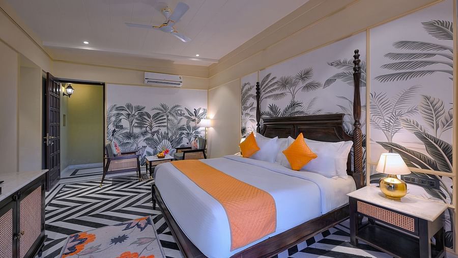 Superior Cottage featuring a four-poster bed with orange accents, chevron-patterned flooring, tropical wallpaper, a seating area, and a nightstand with a gold lamp - Singhvilas, Ranthambore