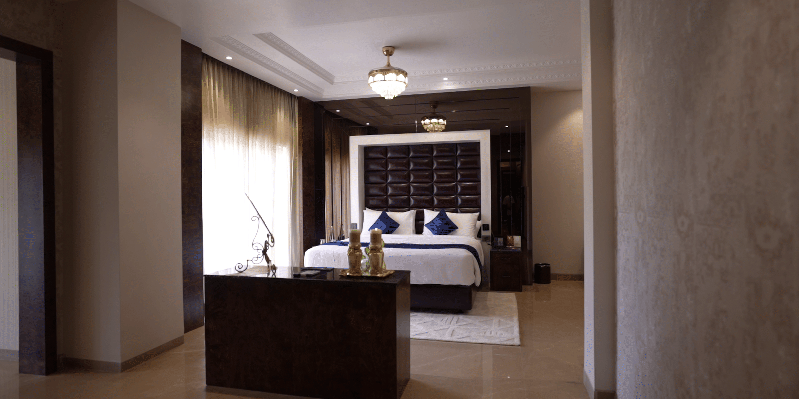 Luxurious bedroom interior with a wooden TV console, large bed with a dark headboard, and natural light from tall windows.