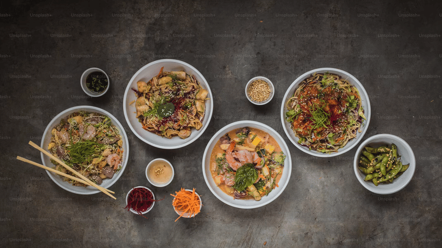 Overhead view of 4 bowls of Asian dishes, including noodle stir-fries, a shrimp curry, and edamame, with chopsticks and small bowls of condiments.