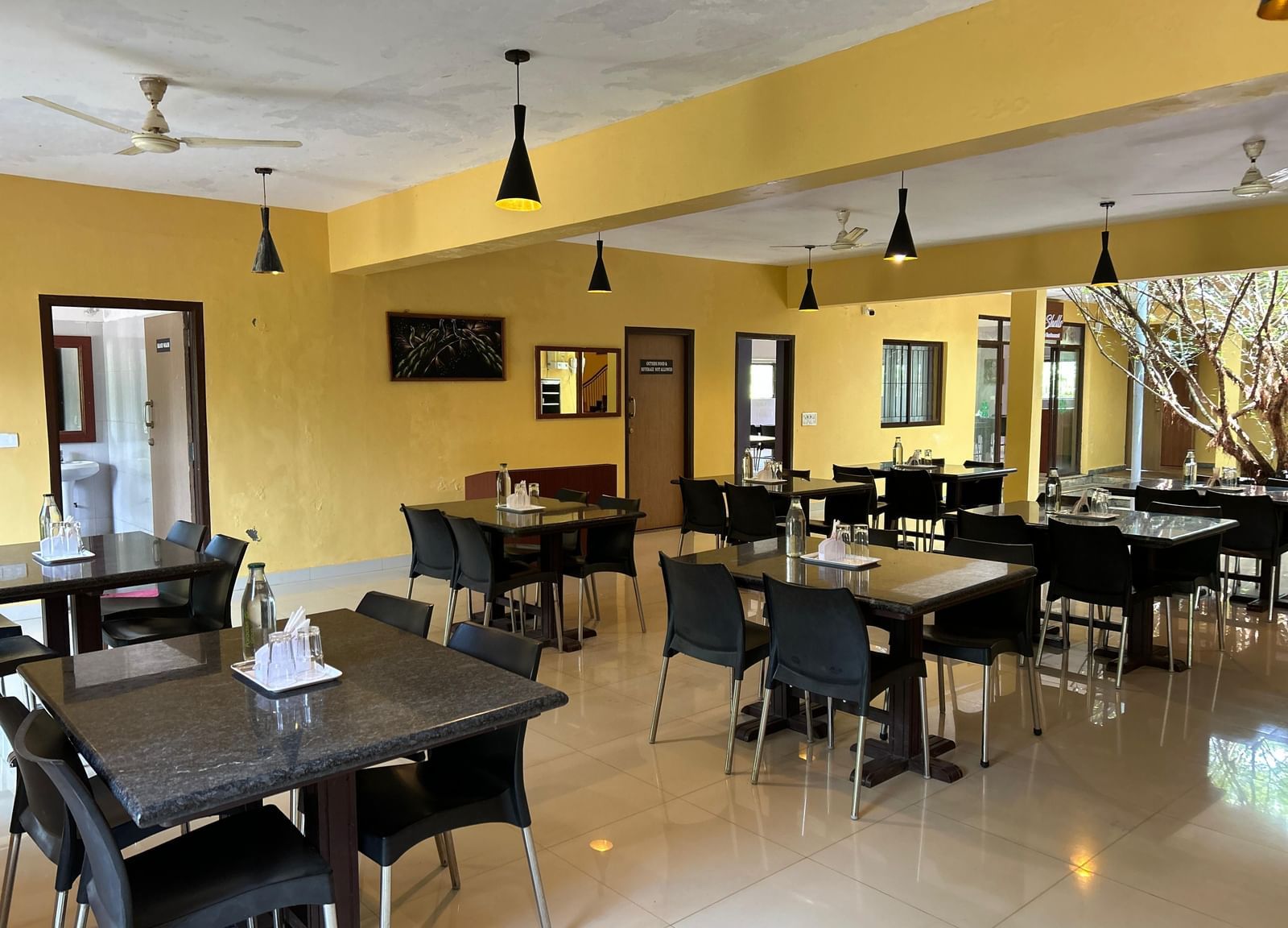 Dining room with yellow walls, tables, black chairs, and hanging lights at Paradise Wild Hills Resort, Kollur.