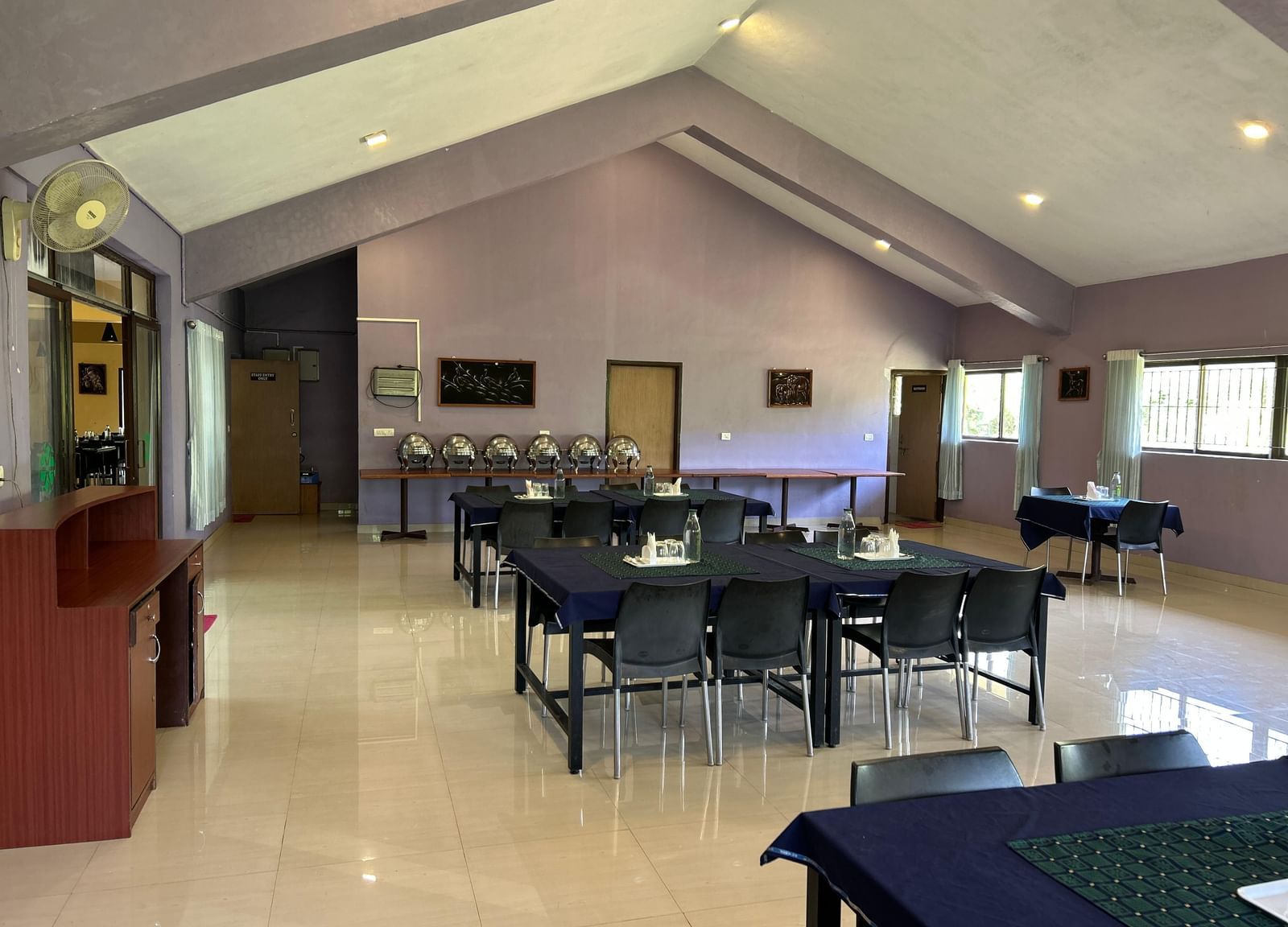 Indoor dining setup with tables, chairs, light purple walls, and ceiling lights at Paradise Wild Hills Resort, Kollur.
