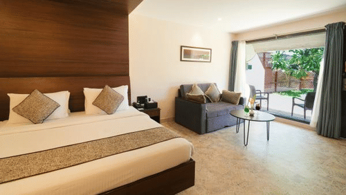 A well-lit hotel room features a king-size bed with neutral linens, a gray loveseat, a round glass table, and a balcony overlooking greenery - MGM Mark Whitefield