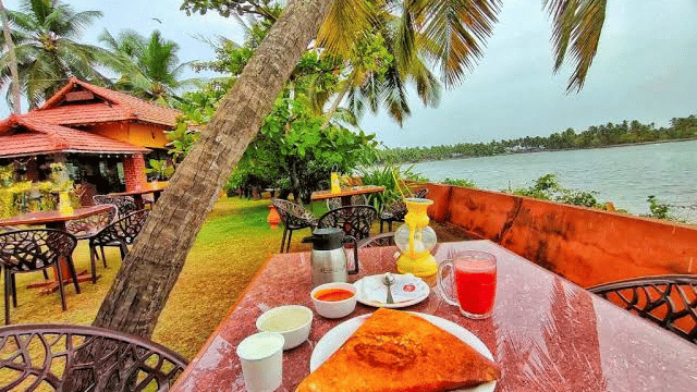 An outdoor dining table set with breakfast items including dosa, juice, and tea, overlooking a river lined with palm trees at Paradise Lagoon Resort, Udupi.