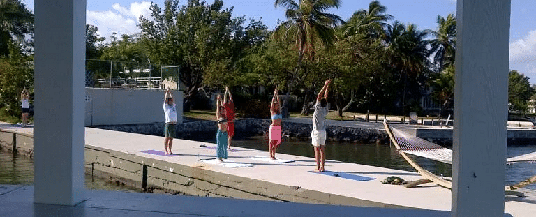 Outdoor view from Casa Morada showing a yoga session on a dock with palm trees and a clear blue sky.