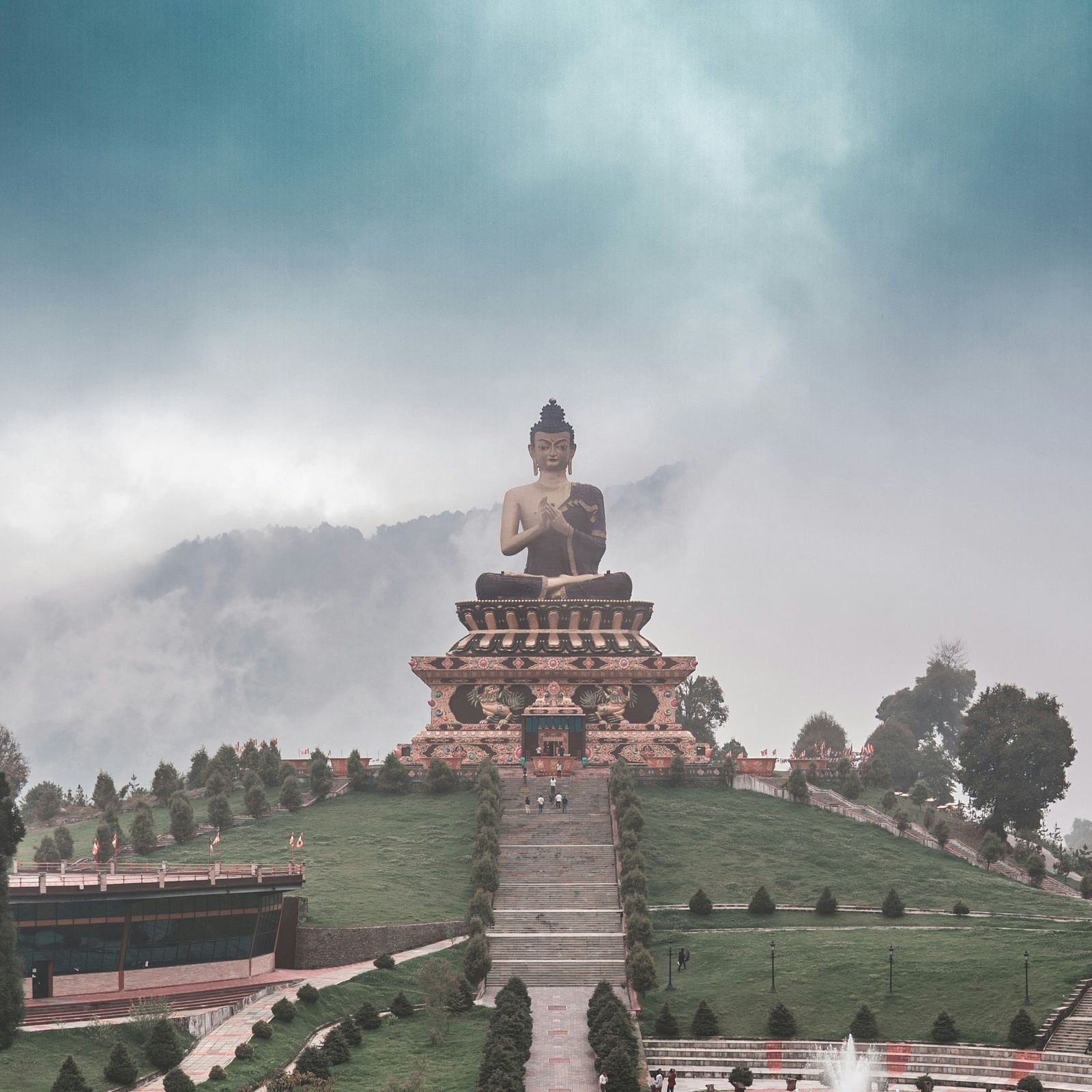 A majestic statue of Buddha atop a hill with a scenic backdrop of cloudy skies and lush greenery.