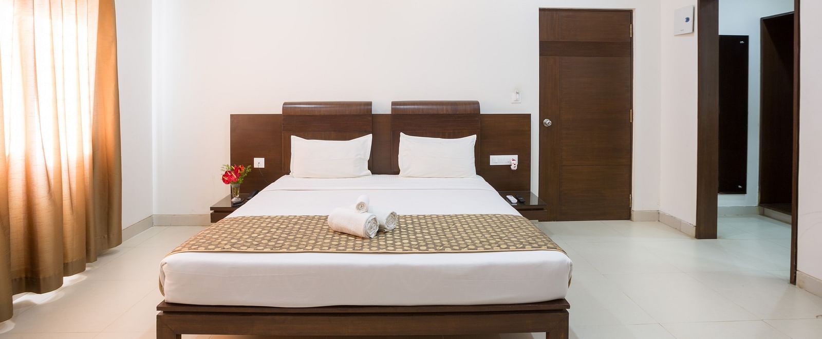 A spacious room with a double bed, dark wooden furniture, and a television on the wall - Sanctum Suites BEL Road, Bangalore