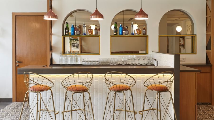 A bar counter at Monday Hotels Mahape, Navi Mumbai, featuring modern bar stools, pendant lights, and arched shelving with bottles.