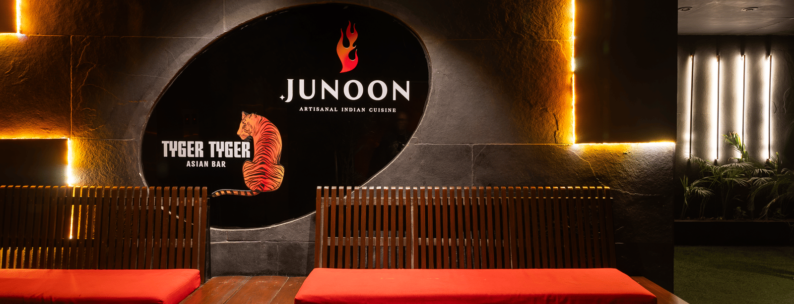 A restaurant entrance with a signboard reading 'JUNOON' above a bench and yellow lights on the wall at Infiniti Hotel, Indore.