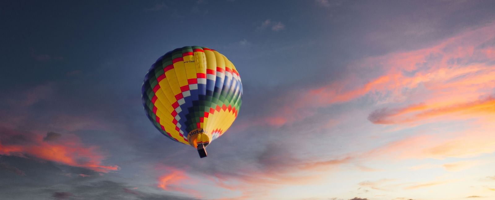 A hot air balloon in view from below with different hues of colours on the clouds above.