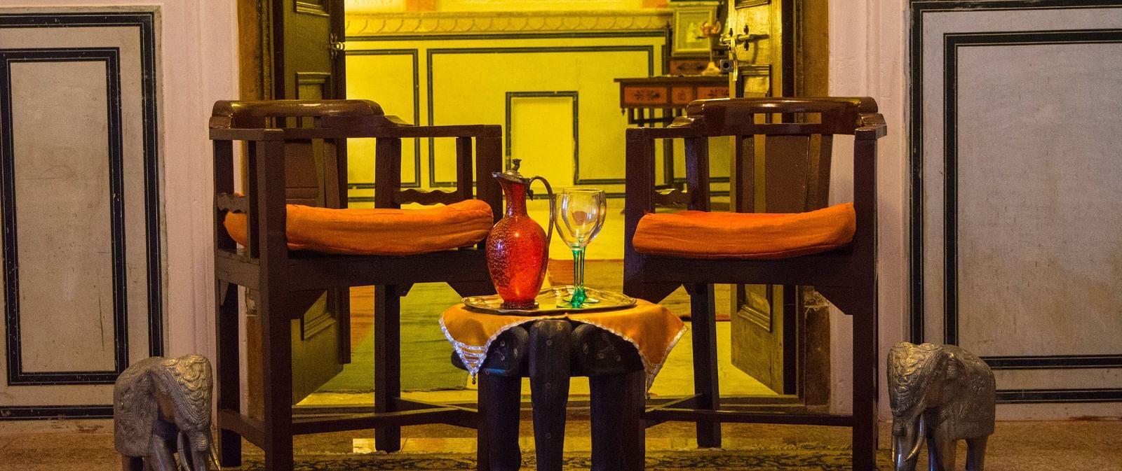 A seating area at Umaid Vilas Jaipur with two wooden chairs, orange cushions, a small table with a red bottle and glasses, and elephant sculptures on either side.