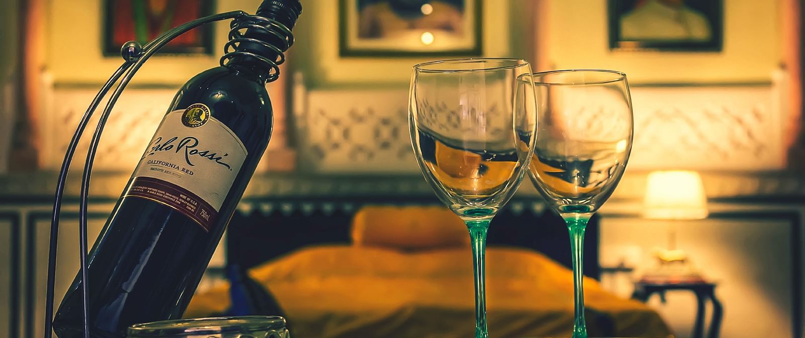 A bottle of wine and two glasses set on a tray in a room at Umaid Vilas Jaipur, with artwork visible in the background.