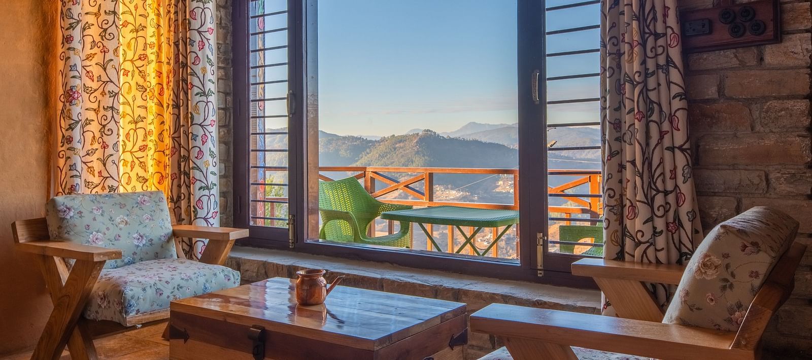 A tranquil interior view showcasing floral curtains, wooden furniture, and a stunning outdoor mountain landscape. - Blusalzz Homes The Himalayan Bungalow, Almora