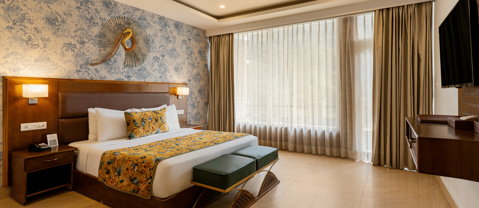 A spacious bedroom featuring a neatly made bed, 2 side cabinets, a wall with blue wallpaper, a decorative ceiling, and sheer and opaque curtains | Summit by the Ganges Beach Resort & Spa, Rishikesh