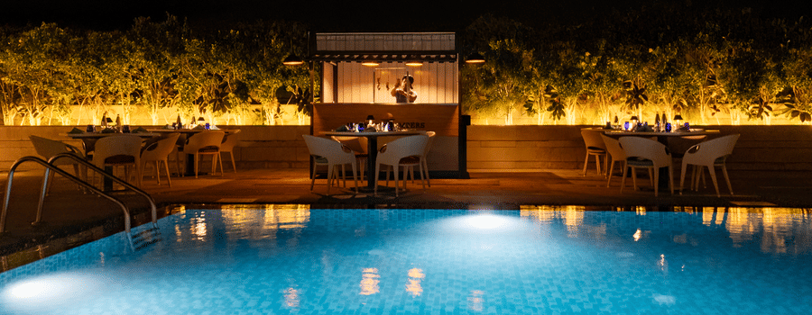 A tranquil nighttime view of the hotel's outdoor swimming pool, lit up with glowing lights, with a seating area surrounded by plants at The Residency Towers Rameswaram.