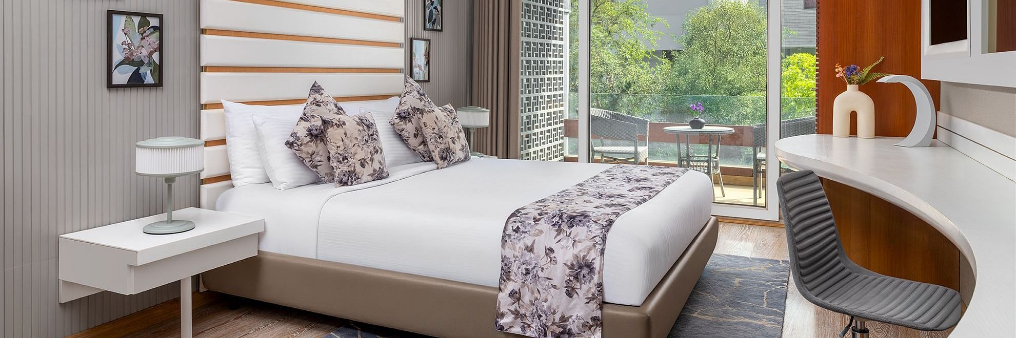 A modern bedroom at The Atara, Gurugram featuring a neatly made bed with a floral runner, a stylish white desk with an armchair, and a large window offering a refreshing view of lush greenery