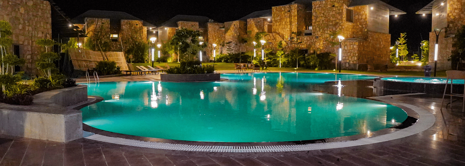 An illuminated swimming pool glows green and reflects the resort buildings at night. The surrounding area is dark with a view of the mountains.