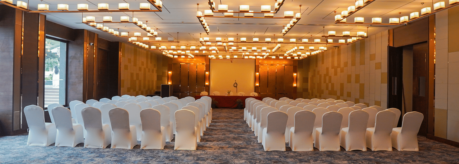 A wide shot of a conference room or ballroom set up with rows of white chairs facing a stage, featuring warm, modern lighting fixtures.