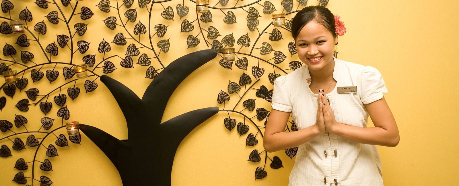  A lady greeting guests in front of a wall which has a tree poster at Grand Chennai By GRT Hotels.