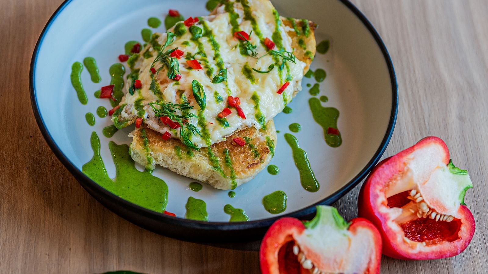 A plate of egg toast topped with sauces and veggies, with capsicum cut into two pieces and placed beside the plate, along with a chilli at Indie Stays Mumbai International Airport.