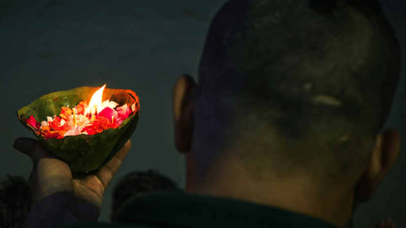 The back of a person's head is visible as they hold a small, lit lamp with flower petals in a dark environment.