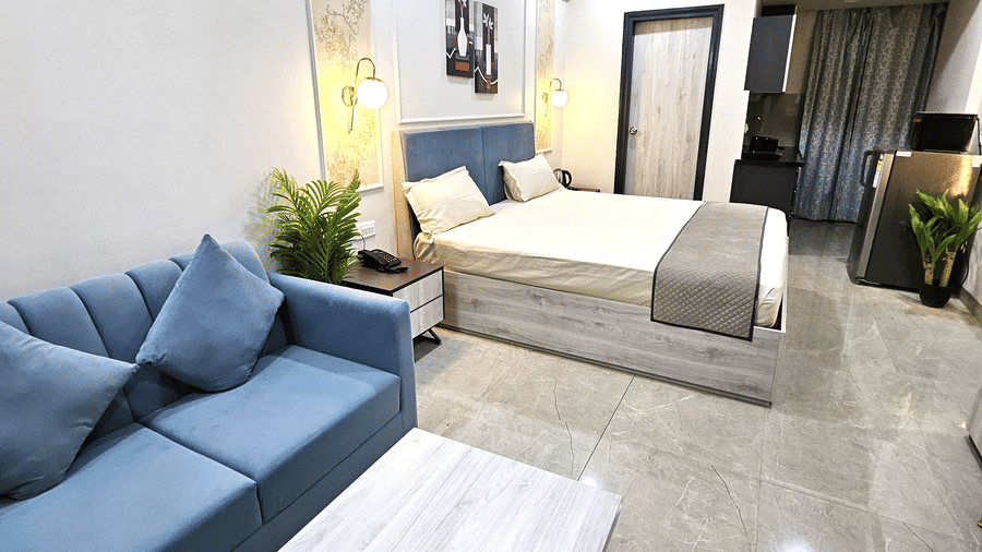 A modern studio room featuring a bed, blue sofa with matching cushions, wooden coffee table, kitchenette with appliances, and warm lighting accents - Urban Suites by BluSalzz, Jaipur