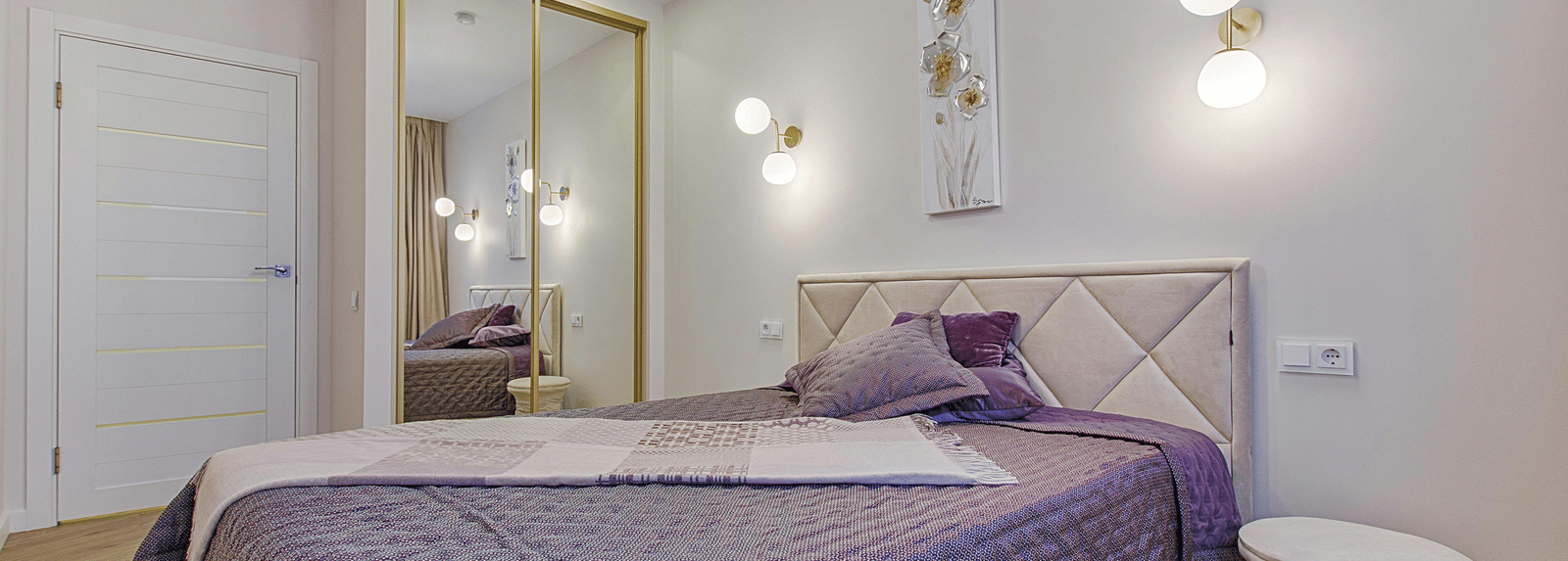 A minimalist bedroom with a quilted purple bedspread, a diamond tufted headboard, and mirrored wardrobe doors.