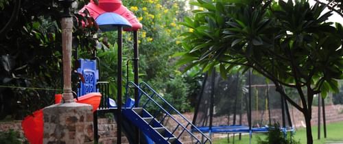 A colorful children's play area with a slide and a trampoline is situated amongst the greenery at Tijara Fort-Palace - 19th Century, Alwar.