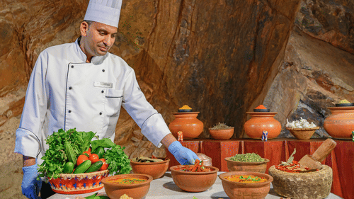 A chef in uniform reaches toward clay pots filled with traditional dishes, fresh vegetables, and spices set against a stone wall | Neemrana Hotels
