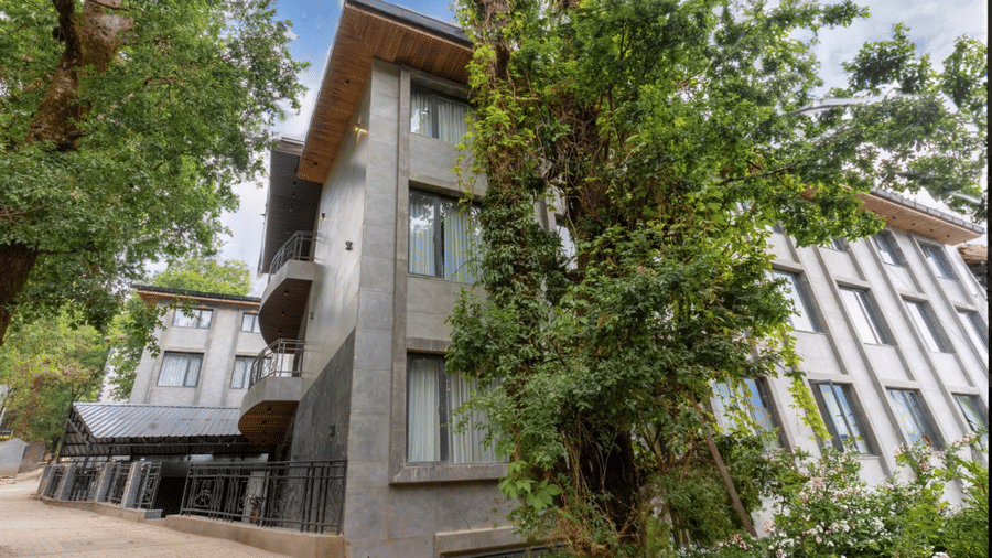 A daylight exterior view of a modern multi-storey grey building surrounded by tall green trees | Lawrence Solitaire Resort, Nainital