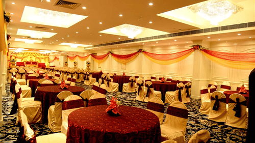 A spacious, well-lit banquet hall with red and gold decor, round tables with red tablecloths, and chairs arranged for a large event - Clarks Avadh, Lucknow